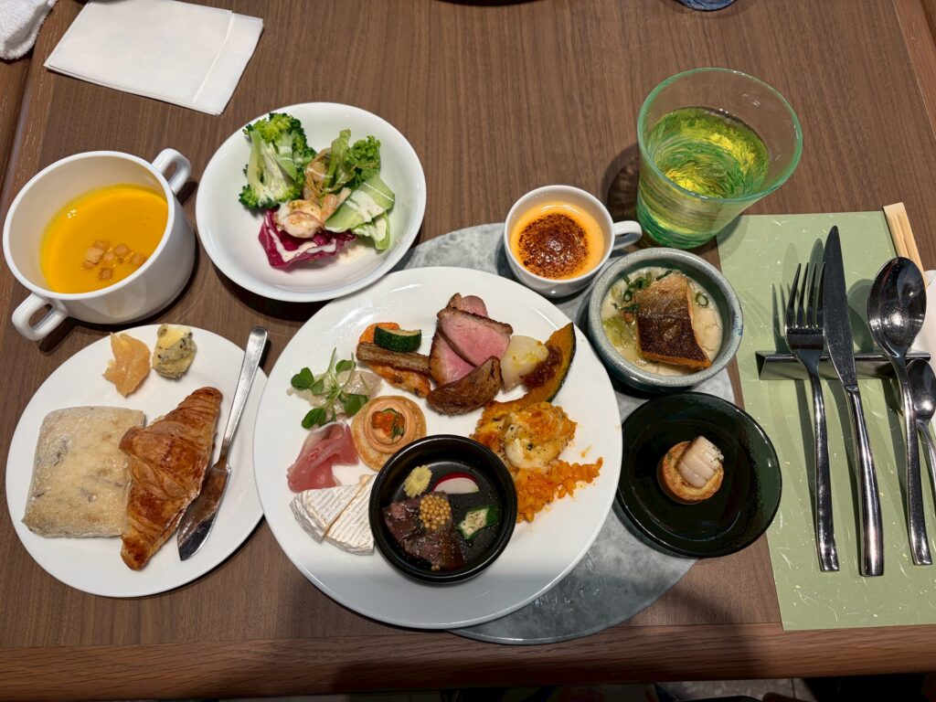 Tabletop meal with multiple plates: soup in a white mug, bread and croissant on a small plate, small salad bowl, and a large shared plate of assorted meats and sides with a dip bowls and small garnishes.