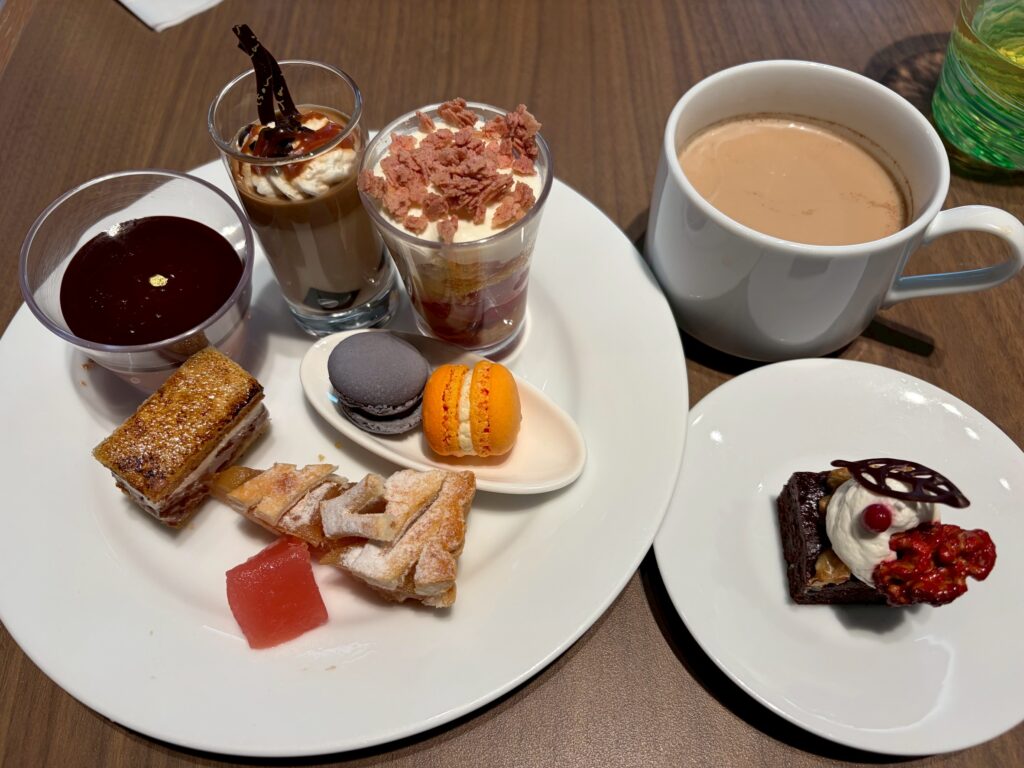 Assorted dessert platter: mini chocolate mousse cups, cream-topped parfaits, flaky pastries, two macarons, a watermelon cube, and a cup of coffee on a wooden table.
