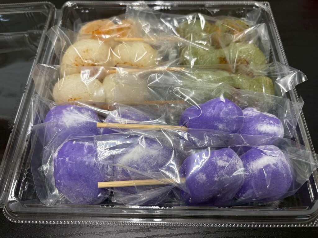 Assorted mochi dumplings in a clear plastic tray, with purple mochi on the bottom and white and green varieties above, wrapped in plastic film.