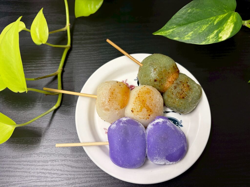 Plate of colorful mochi balls on skewers (purple, white, green, and brown) with green leaves in the background.