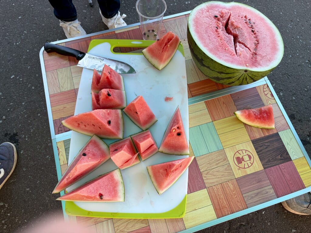 Watermelon pieces arranged on a cutting board with a large halved watermelon on a colorful outdoor table. A knife rests on the board and a plastic cup sits nearby.