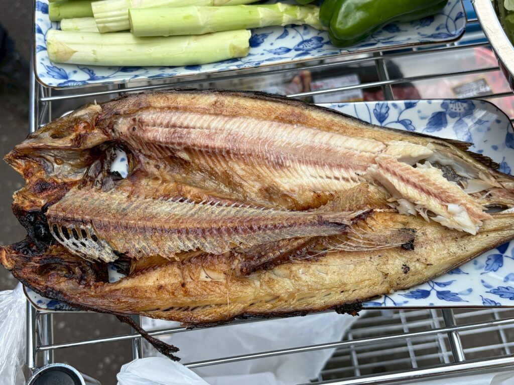 Whole grilled fish served on a decorative blue-and-white plate, crispy skin and flaky white flesh exposed. A bed of celery and a green pepper sit nearby on a rack.