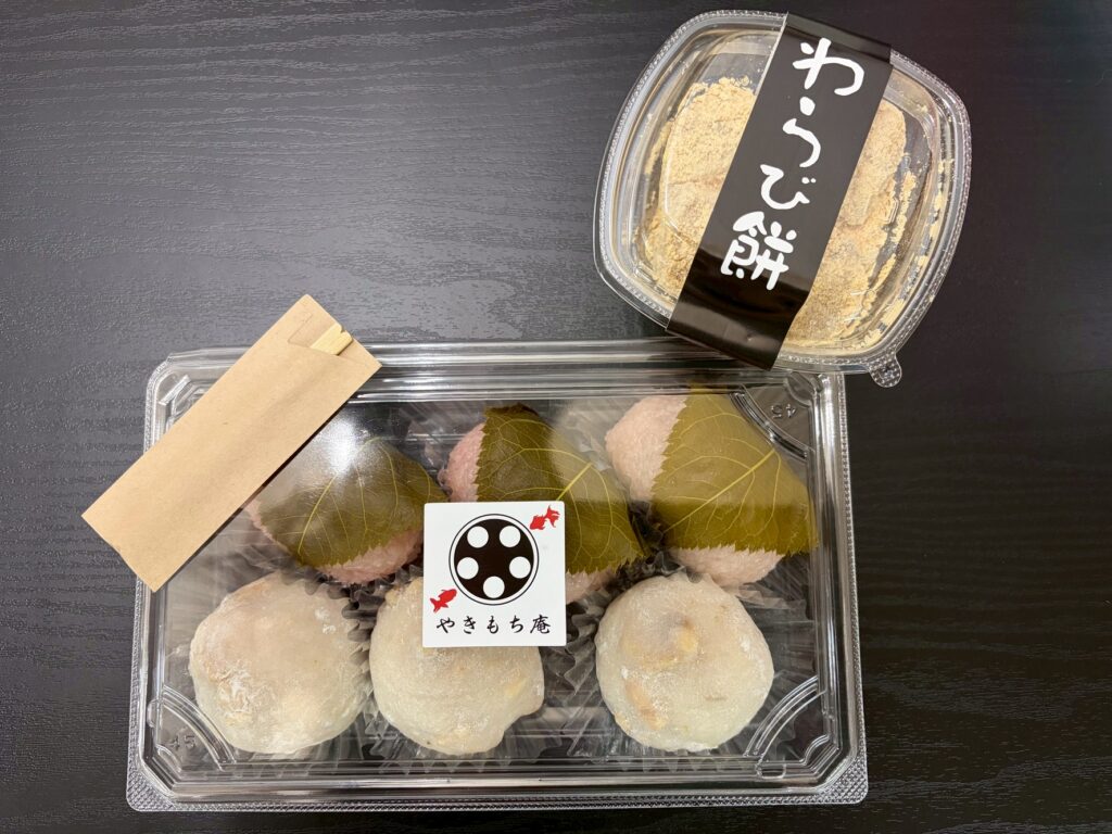line_oa_chat_260728_152238 - 札幌の注文住宅なら寿建設|屋上庭園とガレージが得意な工務店 Leaf-wrapped mochi sweets in clear plastic trays on a dark table, with a black Japanese label on top of one tray.