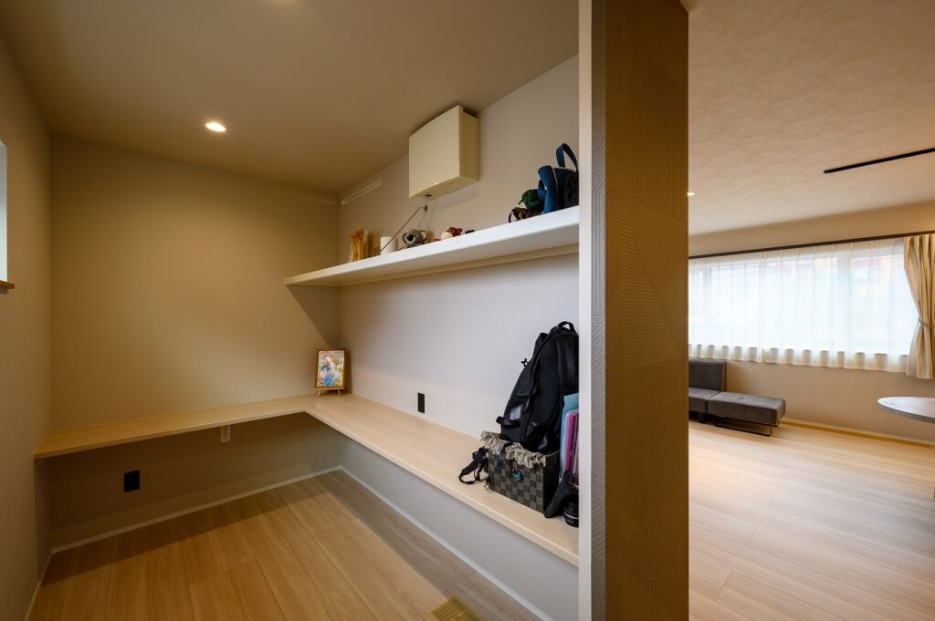 Corner study area with a long wooden desk and wall shelf, a backpack on the desk, and a view into a bright living room.