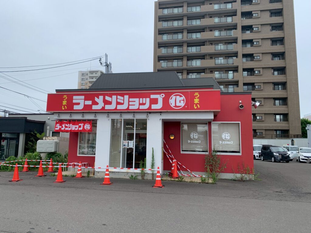 line_oa_chat_260730_171819 - 札幌の注文住宅なら寿建設|屋上庭園とガレージが得意な工務店 Ramen shop storefront with a large red sign reading ラーメンショップ, orange traffic cones in the parking lot, and a tall apartment building in the background.
